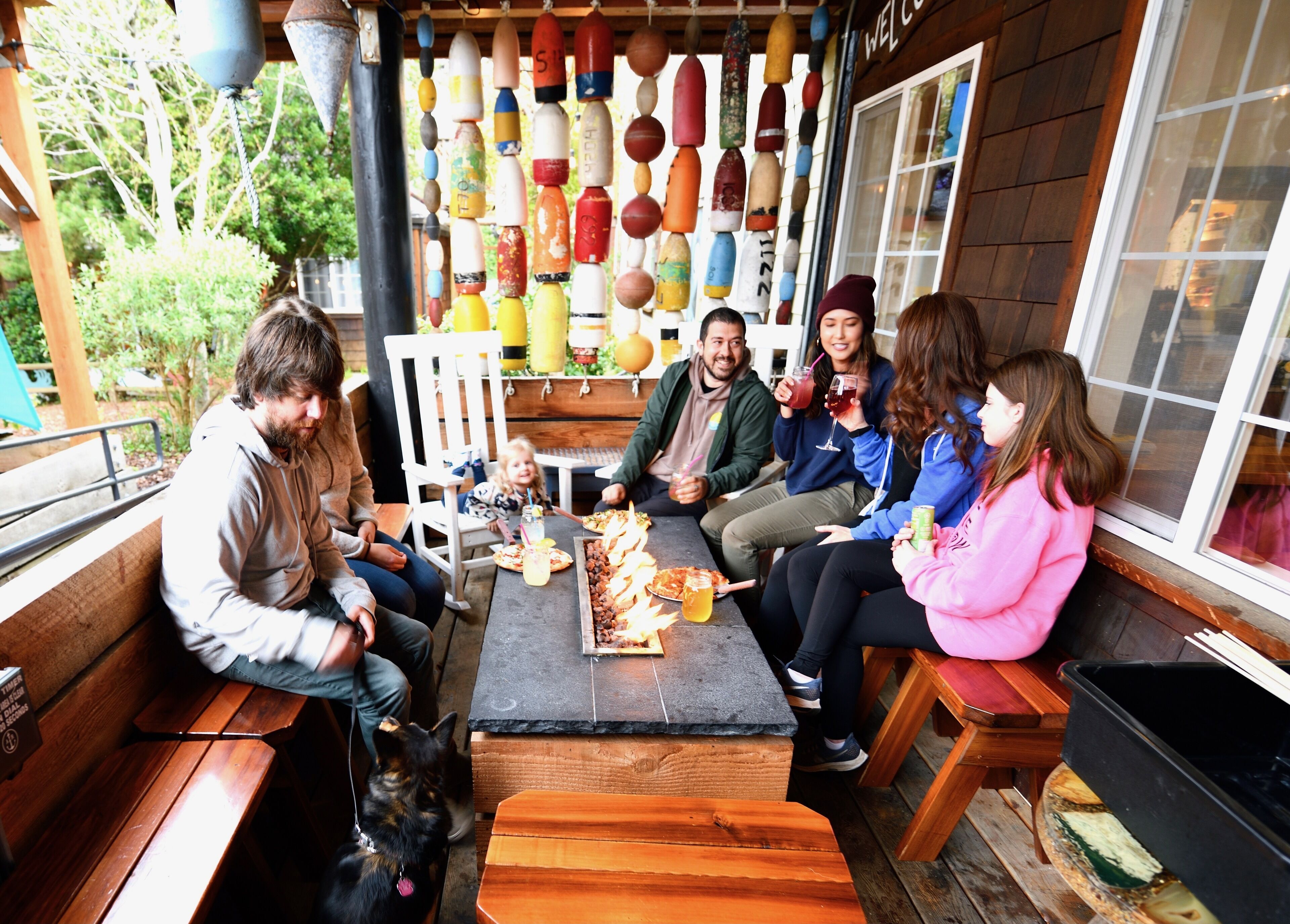 Un grupo de personas sentadas en una mesa de madera con pizza y bebidas, rodeadas por una pared decorada con botellas de colores.