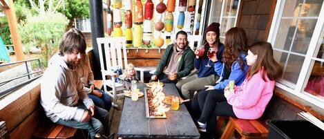 Un grupo de personas sentadas en una mesa de madera con pizza y bebidas, rodeadas por una pared decorada con botellas de colores.