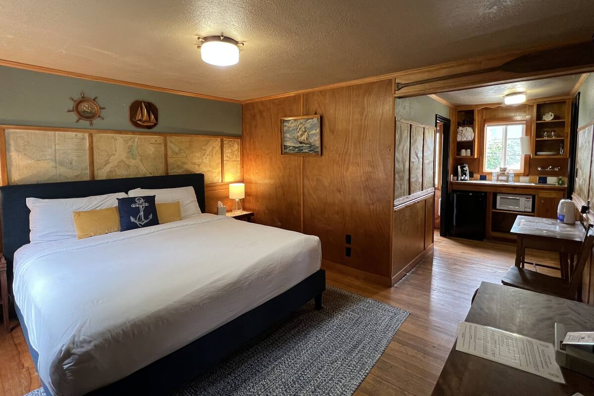 A hotel room with a large bed, wooden headboard, a desk with a chair, a kitchenette, and a view of the outdoors.