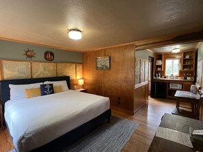 A hotel room with a large bed, wooden headboard, a desk with a chair, a kitchenette, and a view of the outdoors.