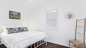 A bedroom with a bed, a window with blinds, a wooden ladder, a wall-mounted mirror, and a tropical-themed painting. - 1-17 Heath Street - Stylish Unit on Heath Street (EVANS HEAD)