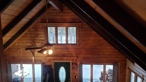 A cozy cabin interior with a sloped ceiling, a chandelier, a dining table, and a view of the outside through large windows. - 10 min from Stratton! Log Cabin w/ Beautiful Views (South Londonderry)