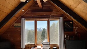 A cozy attic bedroom with a sloped ceiling, a bed with patterned bedding, a desk with a lamp, and a view of a mountain through a window. - 10 min from Stratton! Log Cabin w/ Beautiful Views (South Londonderry)