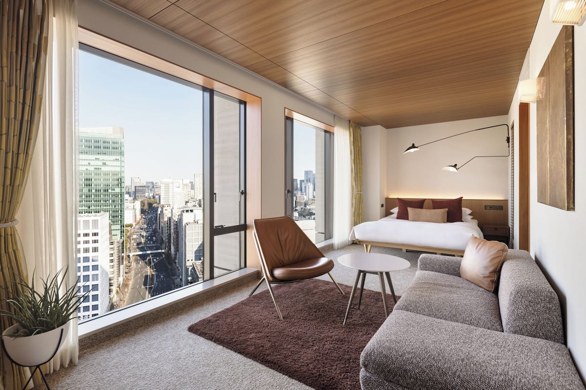 A modern hotel room with a large window, a bed, a sofa, a chair, and a small table.
