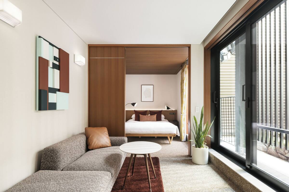 A modern bedroom with a bed, a sofa, a small table, a potted plant, and a sliding door to a balcony.