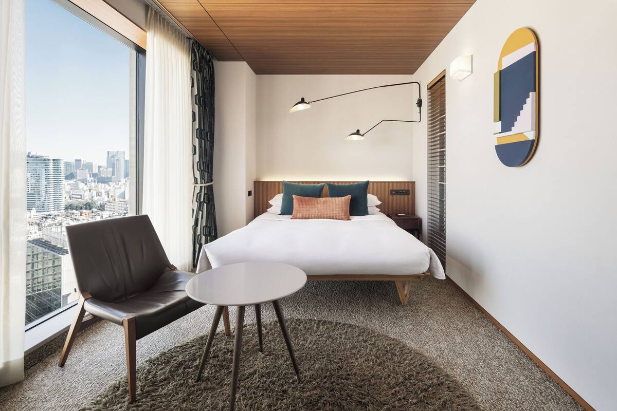 A modern hotel room with a large bed, a view of the city, a chair, a small table, and a unique wall art piece.