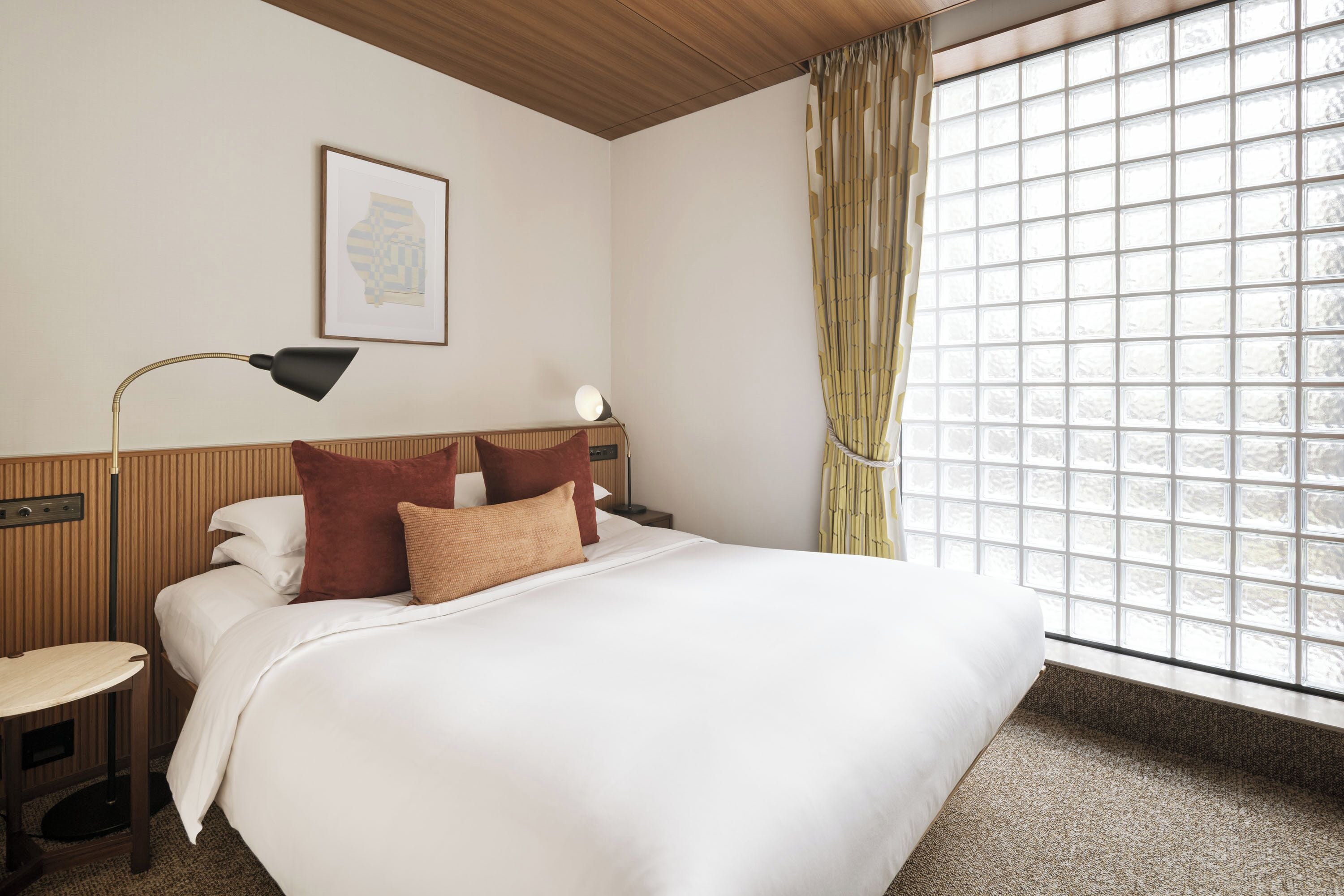 A hotel room with a bed, bedside table, lamp, and window with curtains.