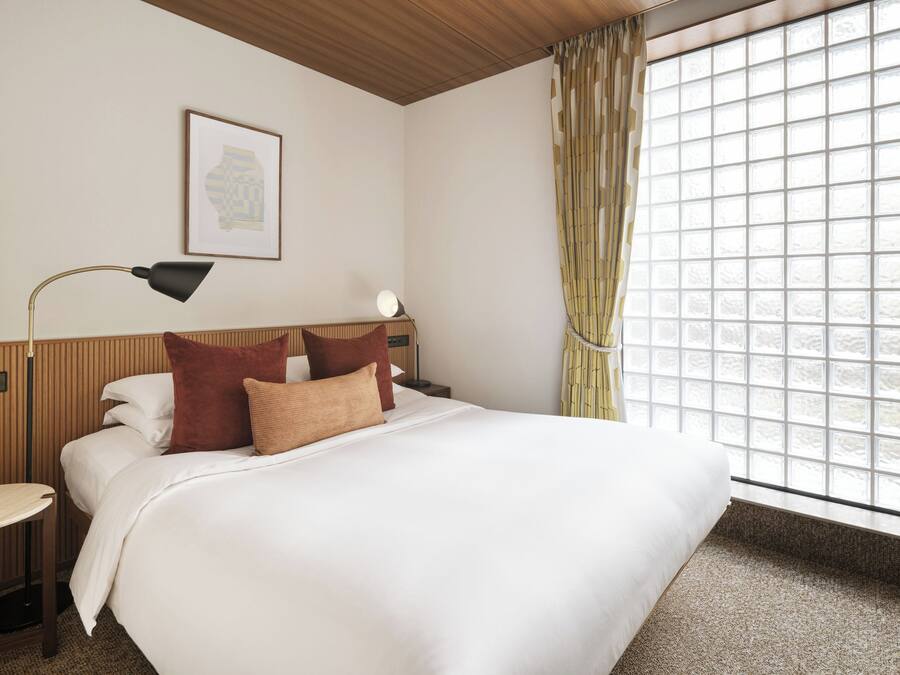 A hotel room with a bed, bedside table, lamp, and window with curtains.