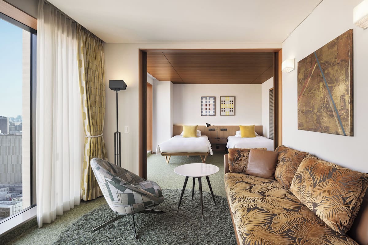 A hotel room with a sofa, armchair, round table, two beds, and artwork on the walls.