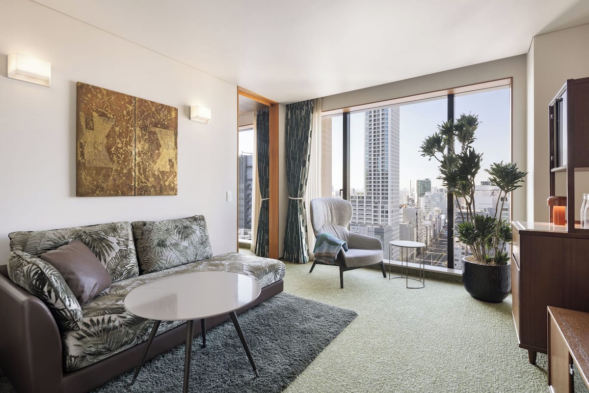 A modern living room with a sofa, armchair, round table, and a large window with a city view.