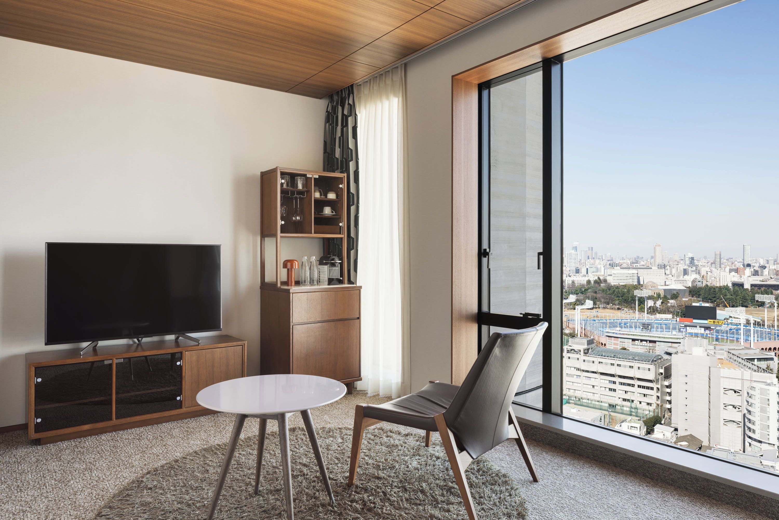 Quarto de hotel moderno com uma TV grande, um armário de madeira, uma mesa redonda, uma cadeira e vista para a cidade através de amplas janelas.