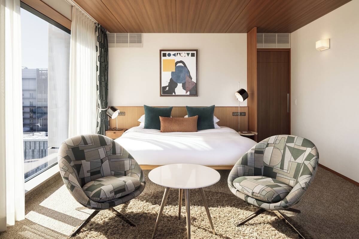 A hotel room with a large bed, two armchairs, a small table, a poster on the wall, and a window with curtains.