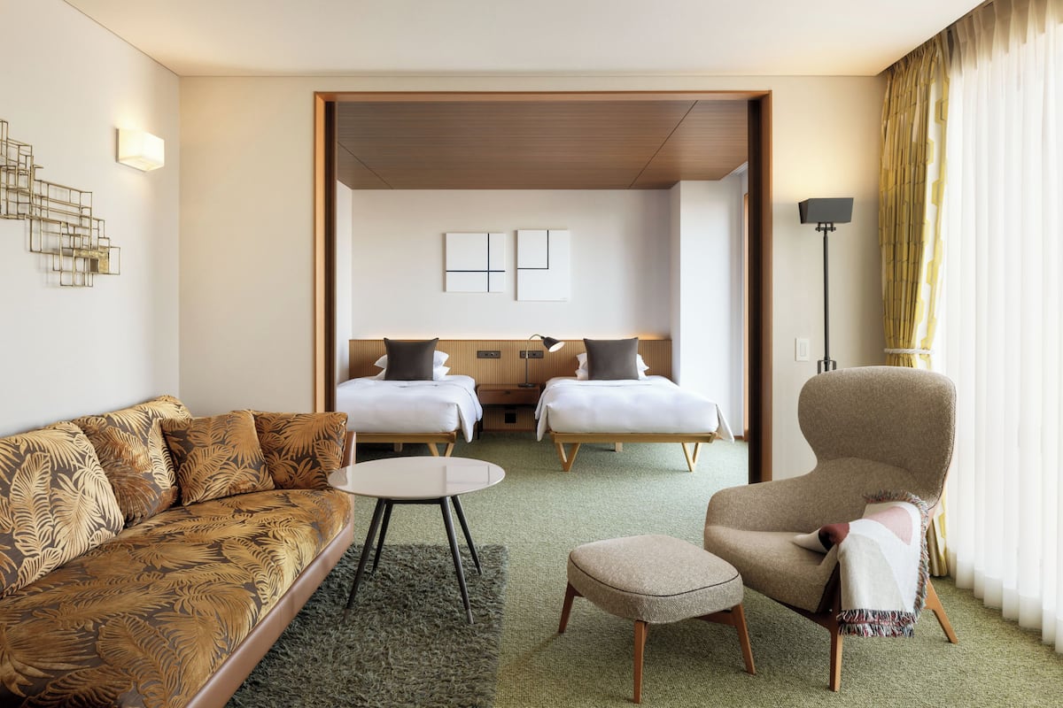 A hotel room with a sofa, two beds, a chair, a small table, and a lamp.