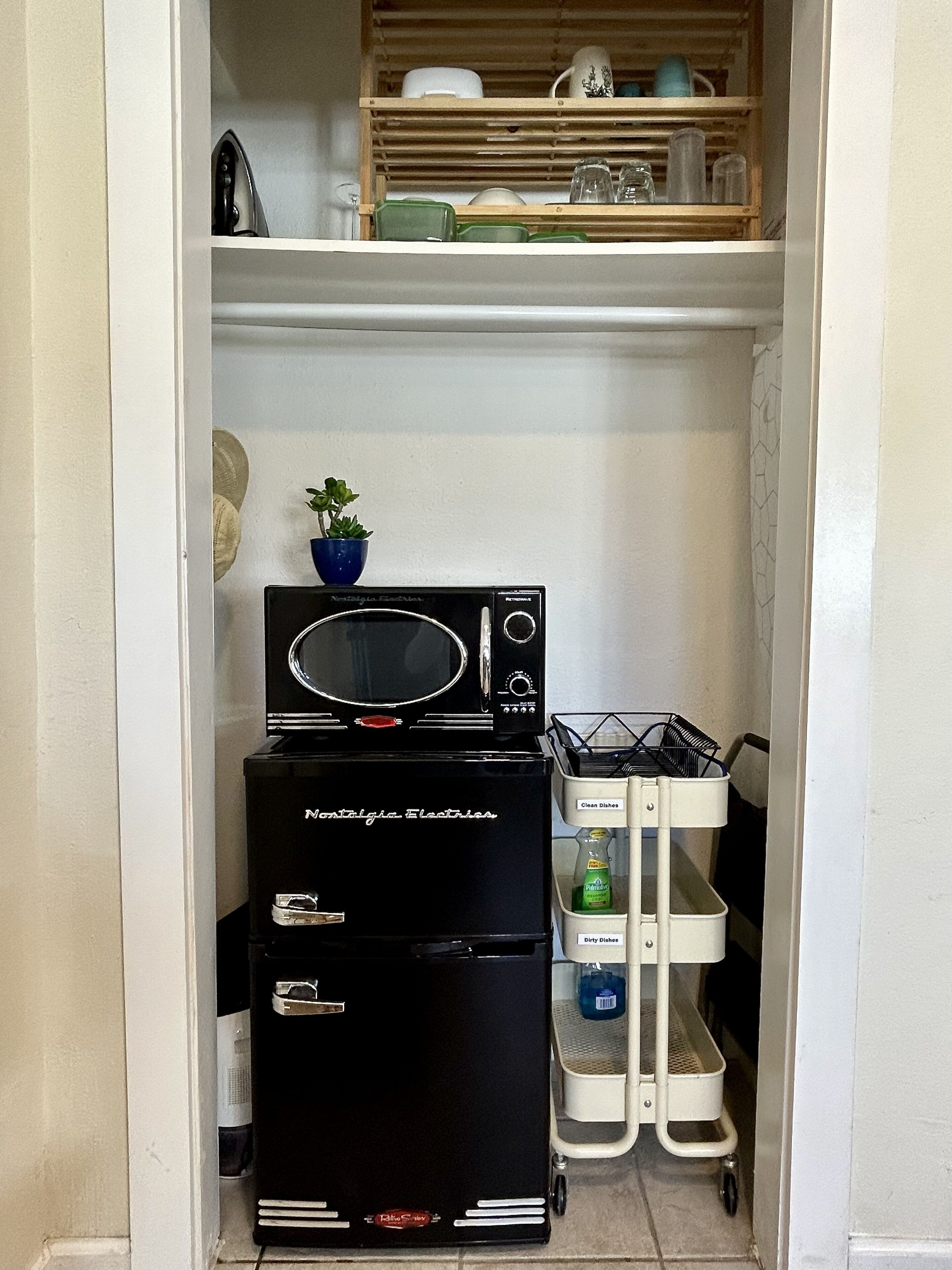 A compact kitchen with a microwave, a refrigerator, and a small shelf with dishes and a potted plant.