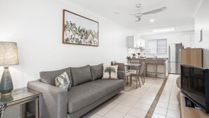 A modern living room with a grey sofa, a dining area, a kitchen with white cabinets, a ceiling fan, and a flat-screen TV. - 1-68 Woodburn Street (EVANS HEAD)