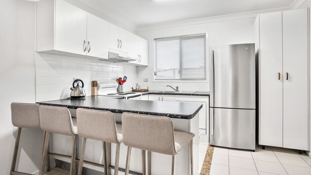 A modern kitchen with white cabinets, a stainless steel refrigerator, a kettle, and a bar area with chairs. - 1-68 Woodburn Street (EVANS HEAD)