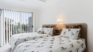 A hotel room with two beds, a nightstand, a lamp, and a view of the outdoors through large windows. - 1-68 Woodburn Street (EVANS HEAD)