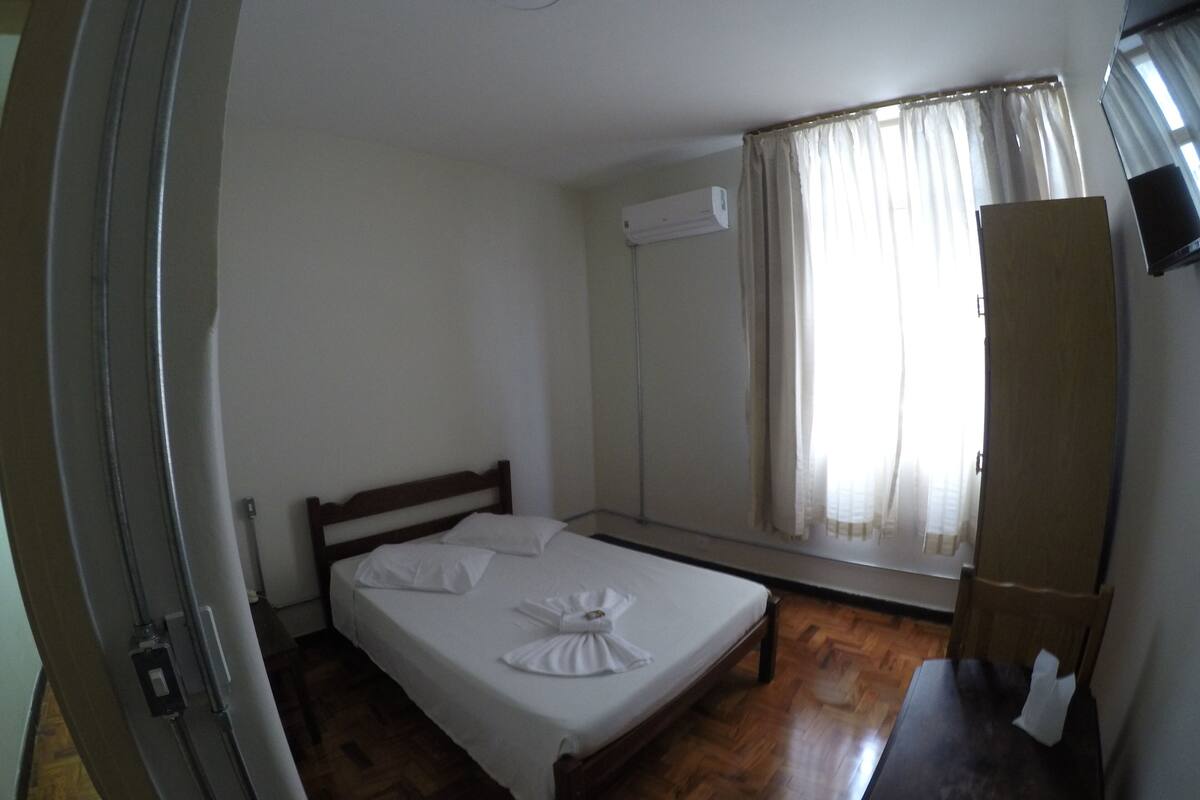 A single bed with a wooden headboard in a room with a wooden floor. There is a mirror on the wall and a window with curtains.