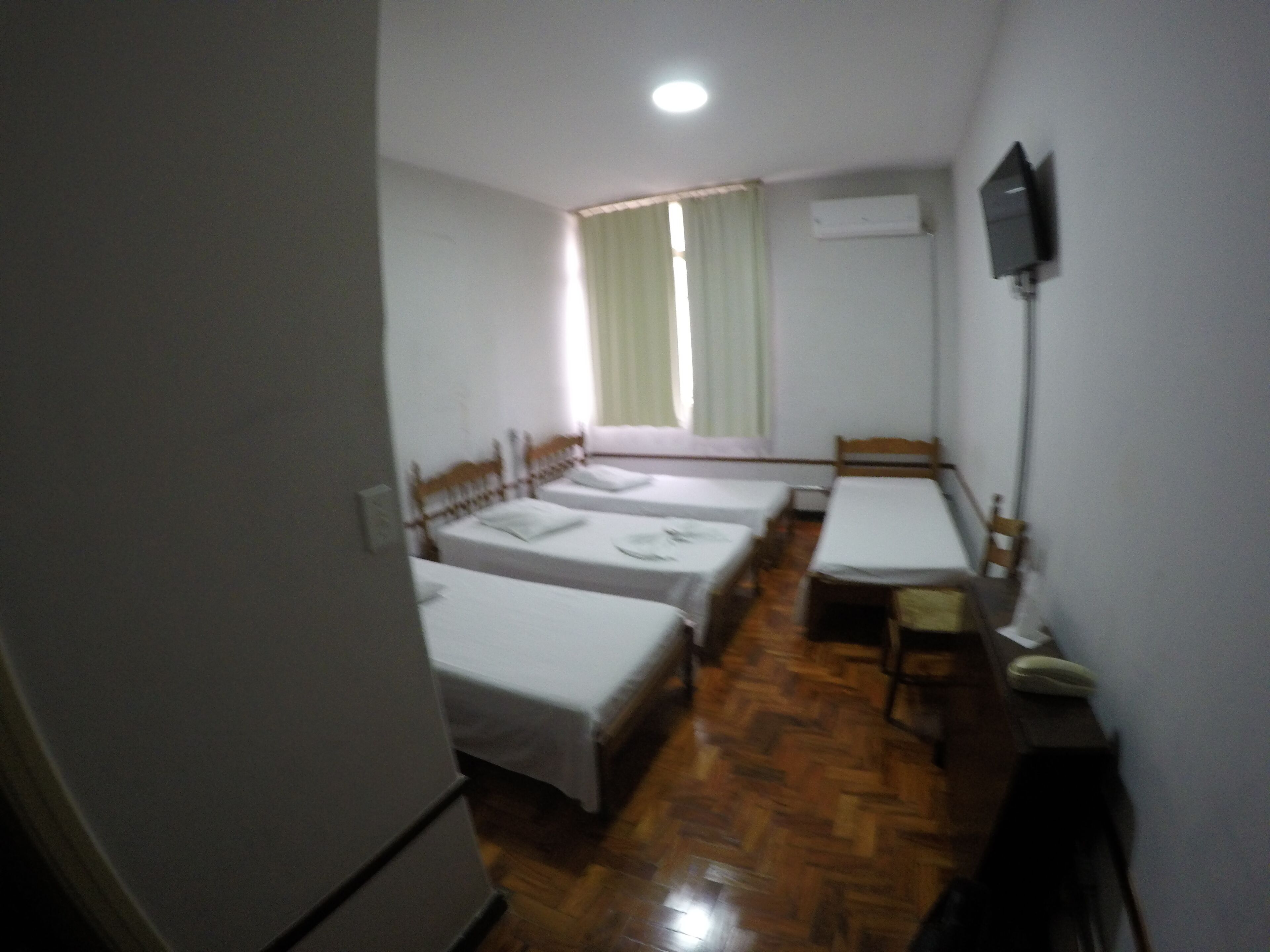 A small, dimly lit room with three single beds, a wooden table, and a television mounted on the wall.