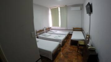 A small, dimly lit room with three single beds, a wooden table, and a television mounted on the wall.