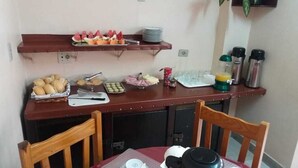 Un pequeño comedor con una mesa dispuesta para dos personas, un estante arriba con platos y salsas, y varios alimentos sobre la mesa.
