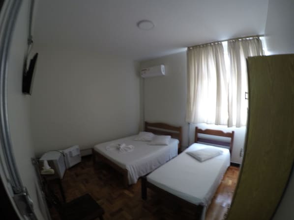 A small, clean room with two single beds, a wooden floor, a window with curtains, and a wall-mounted air conditioner. - Hotel São Nicolau (Taubate)