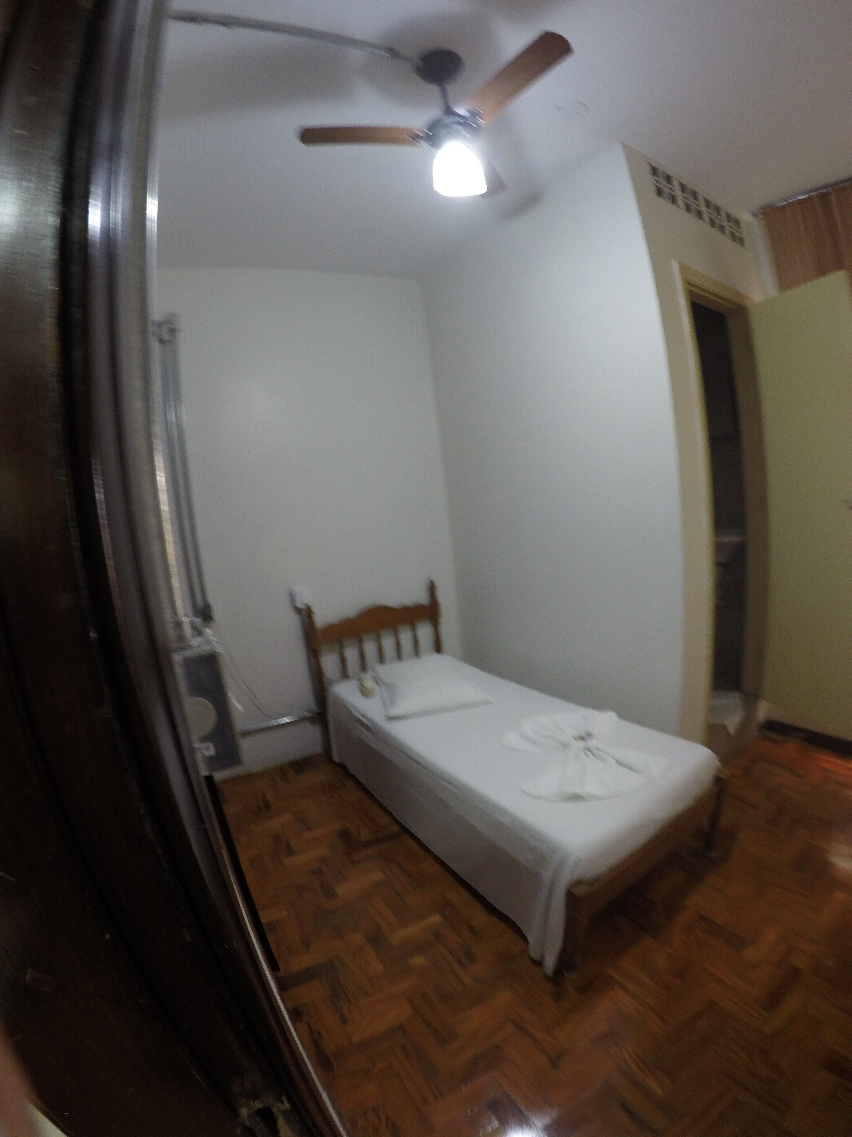 Ein kleines Zimmer mit einem Einzelbett, einem Holz-Kopfteil, einem Deckenventilator und einer Lampe.