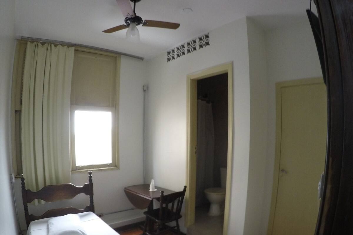 A small, compact room with a single bed, a wooden chair, a small table, and a ceiling fan.