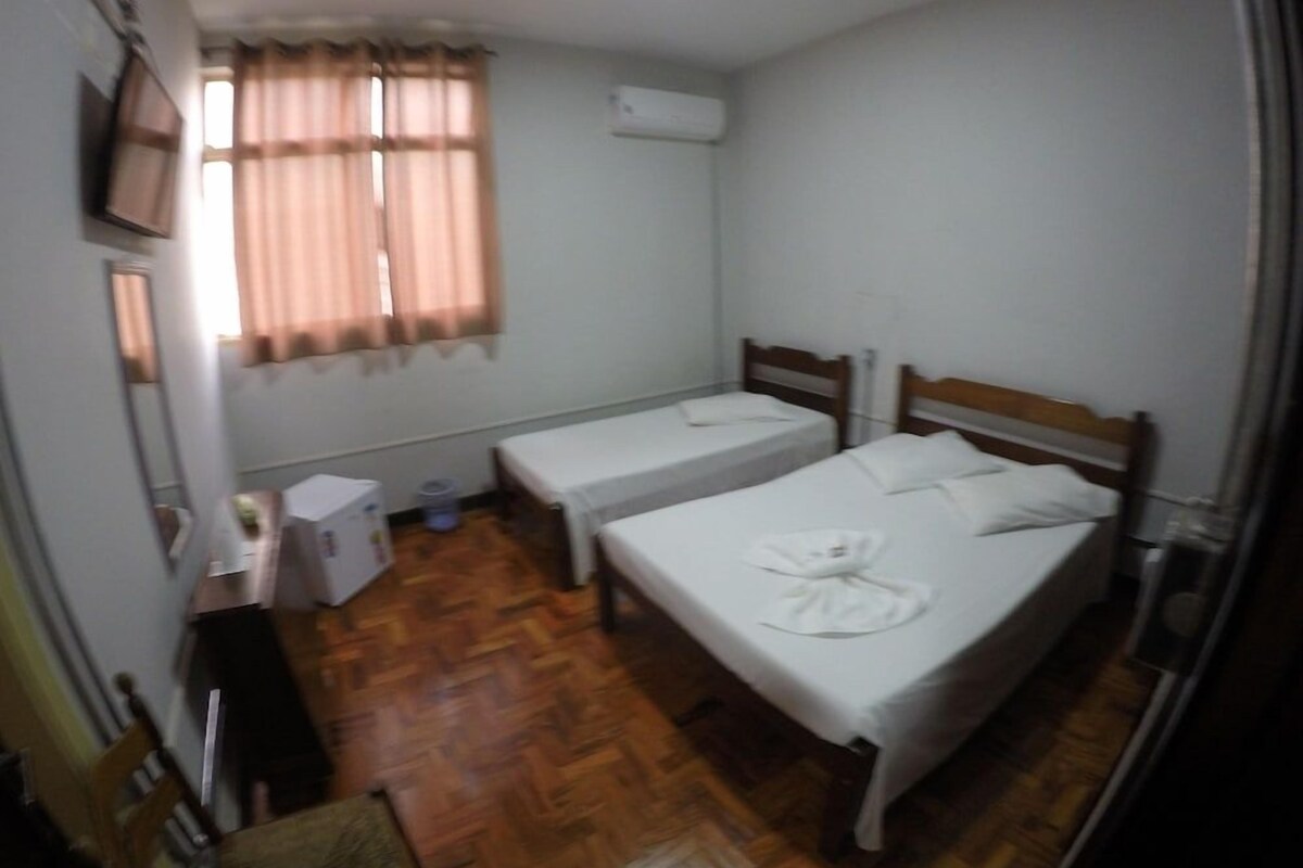 Un alojamiento pequeño y con una sola habitación, con dos camas, piso de madera, un escritorio con silla y una ventana con cortinas.