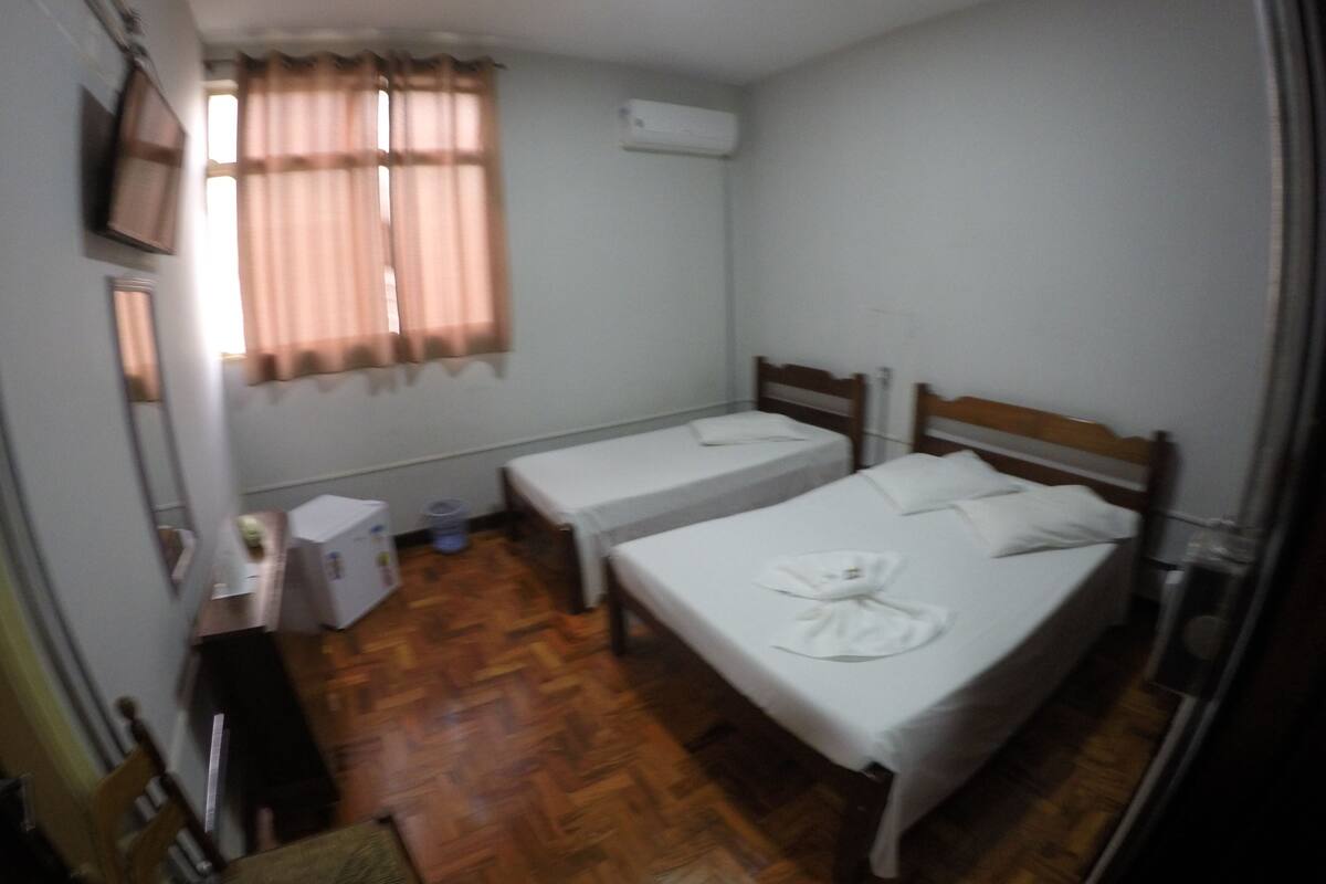 A small, clean room with two single beds, a wooden floor, a TV mounted on the wall, and a window with curtains.