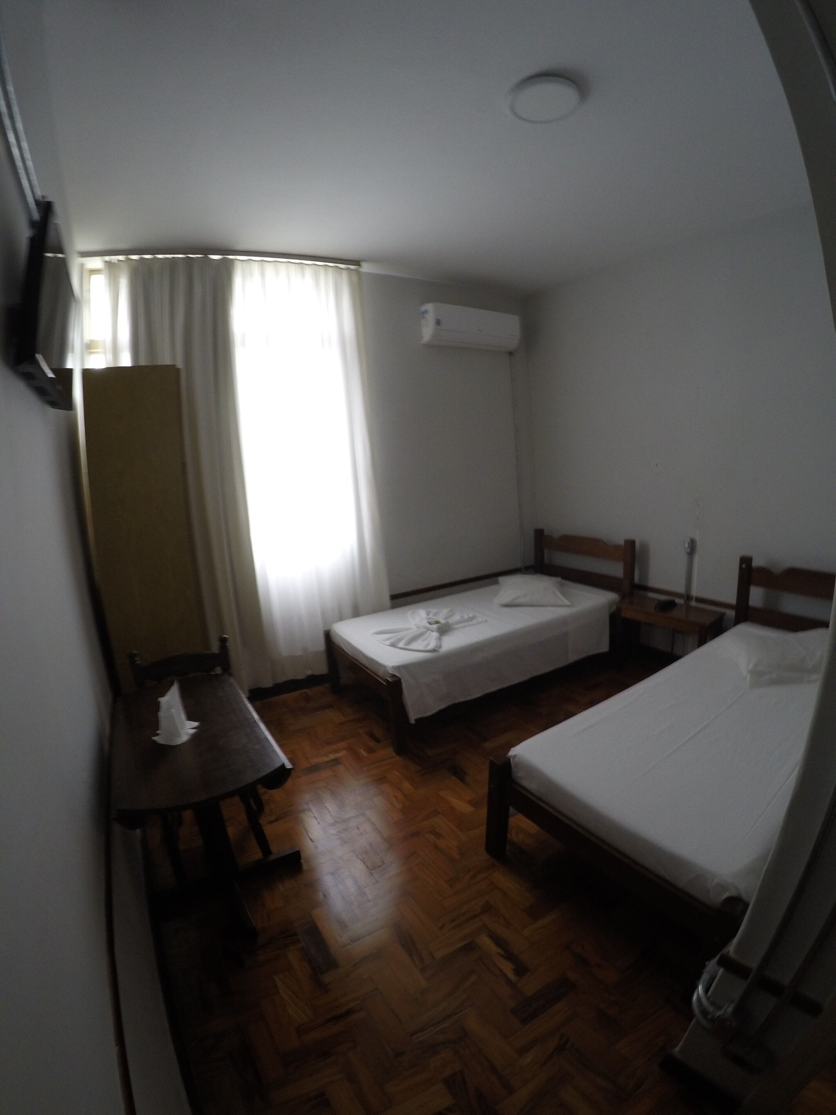 A small, single-room hotel room with a wooden floor, a bed, a bedside table, a chair, a desk, and a window with curtains.