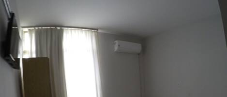 Quarto de hotel pequeno e individual, com piso de madeira, uma cama, mesa de cabeceira, cadeira, escrivaninha e uma janela com cortinas.