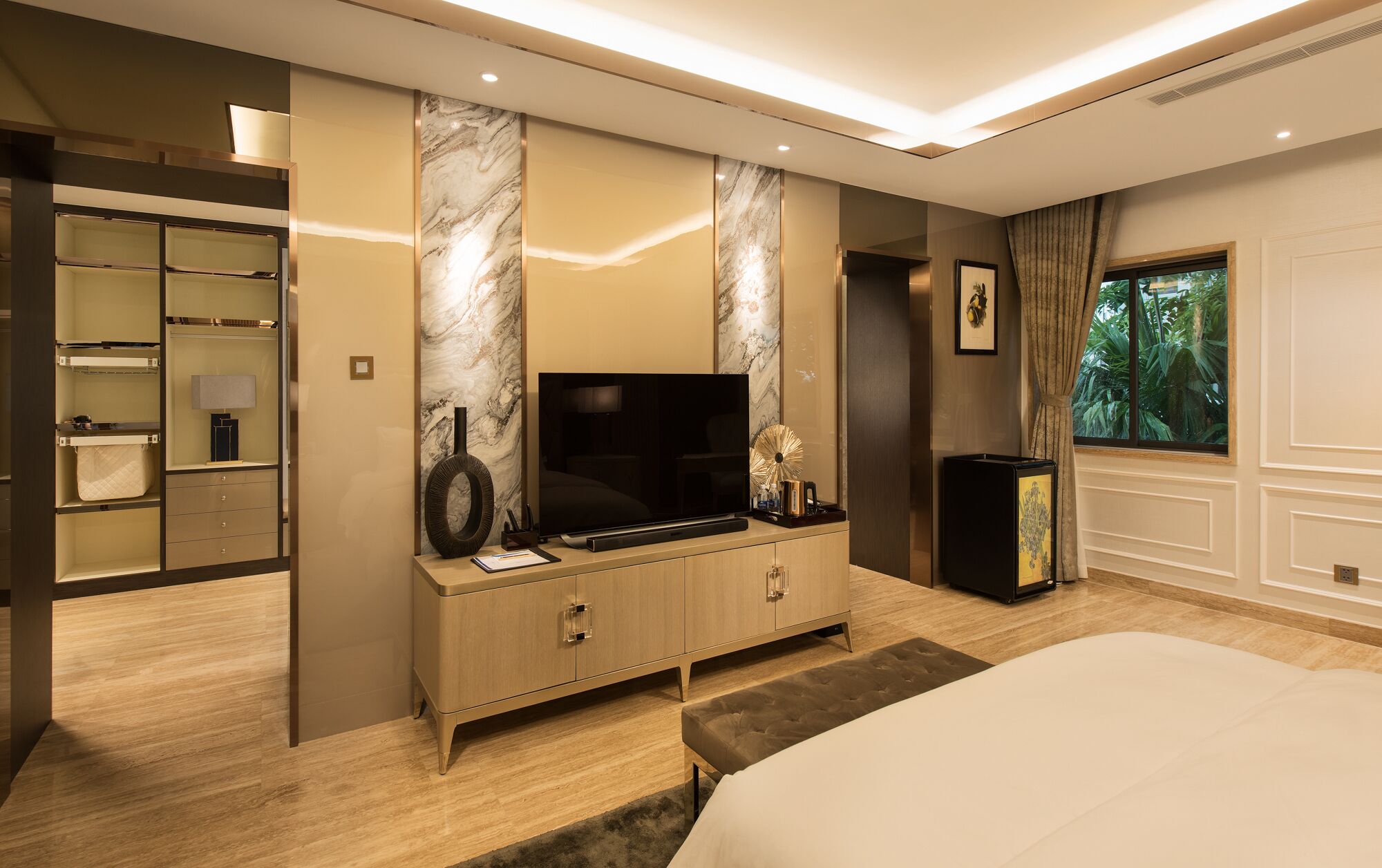 A modern bedroom with a large bed, a TV on a wooden cabinet, a mirror, and a window with a view of greenery.