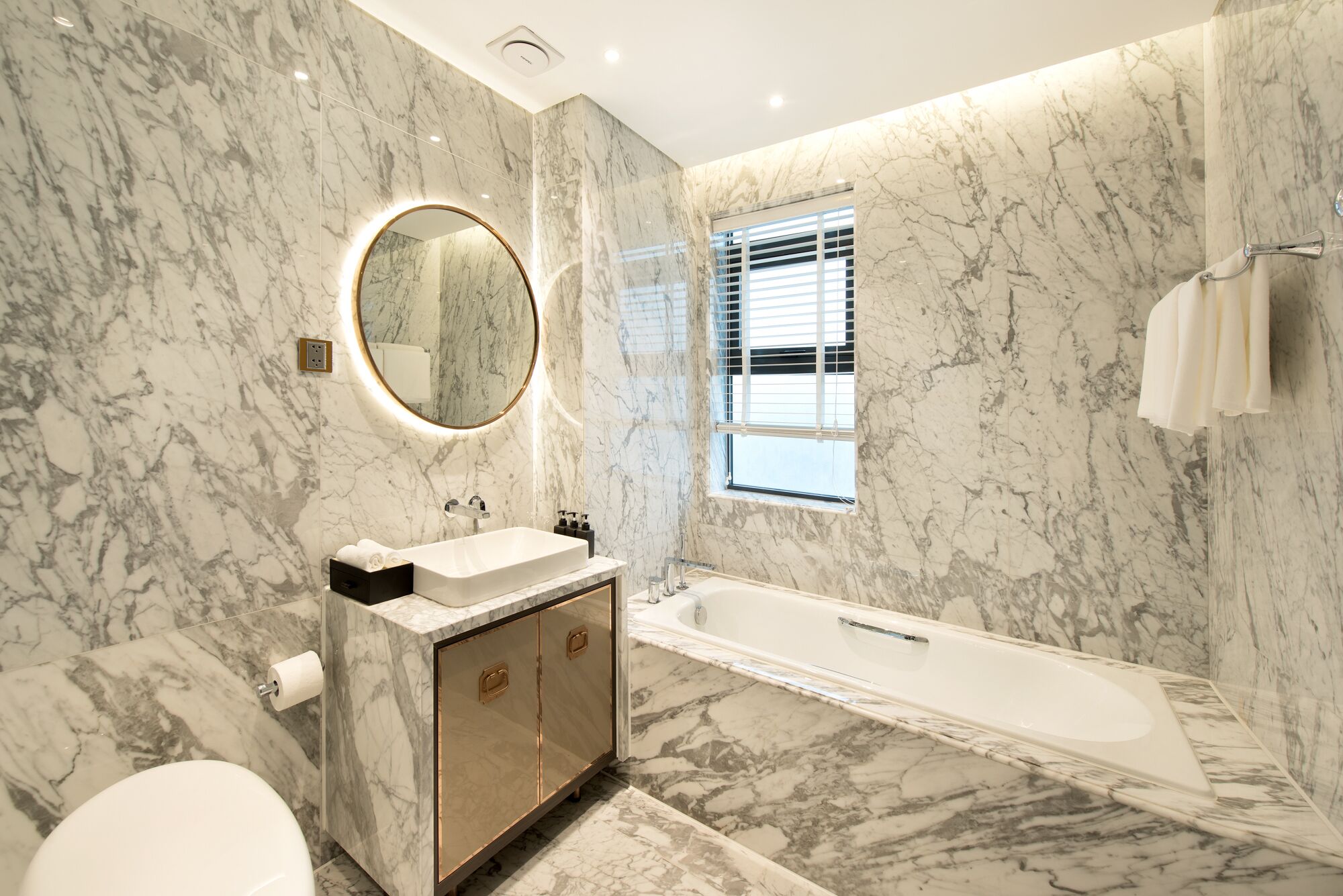 A modern bathroom with a marble floor, a freestanding bathtub, a vanity with a mirror, and a window with blinds.