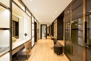 Un pasillo moderno de hotel con suelo de madera, un escritorio con silla, un otoman redondo y armarios con puertas de vidrio.