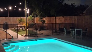 Una piscina con cubierta de madera y cerca. Hay luces colgantes sobre la cubierta.