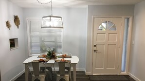 Un comedor con una mesa blanca, cuatro sillas, una lámpara colgante y una puerta con ventana.