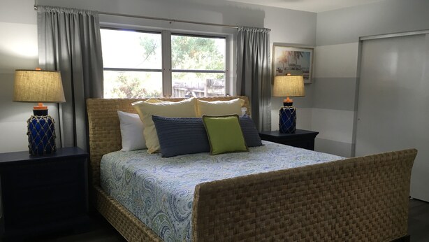 Un dormitorio con cama, dos mesitas de noche, una ventana con cortinas y una puerta.