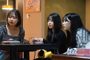 Three individuals seated at a table with electronic devices, in a room with a painting on the wall and a hanging light fixture.