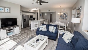 A modern living room with a blue sofa, white coffee table, and a flat-screen TV. There is a kitchen in the background with white cabinets and a dining area with white chairs. - Labor Day special at Celadon 1201: 3 bed/3 bath beachfront (Panama City Beach)