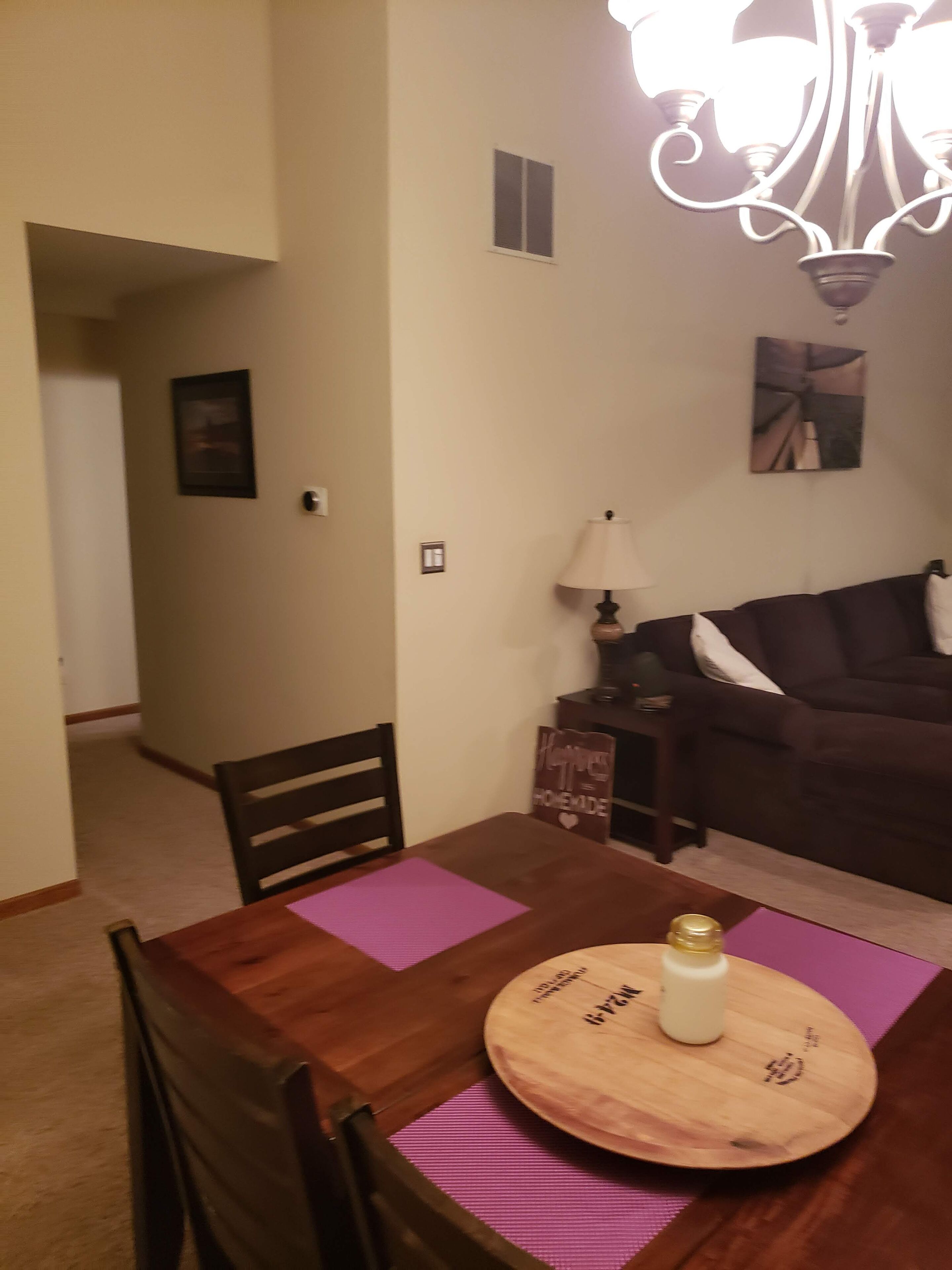 A dining area with a wooden table, purple placemats, and a round wooden board with text. There is a dark brown sofa with white pillows in the background.