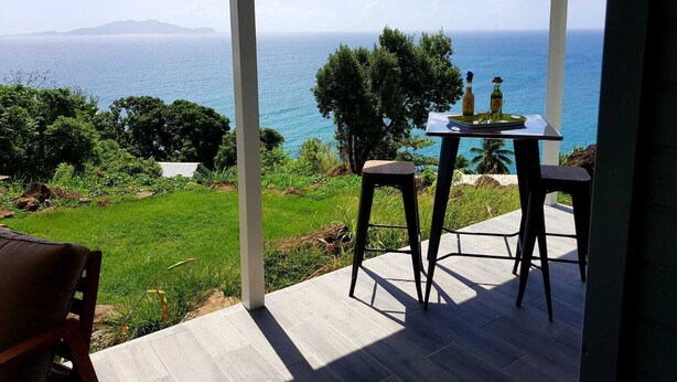 A covered outdoor seating area with a table and chairs, overlooking a scenic view of the sea and distant mountains. - Magnificent views of the sea and islands of Les Saintes, lulled by the sound of the waves. (Trois Rivieres)