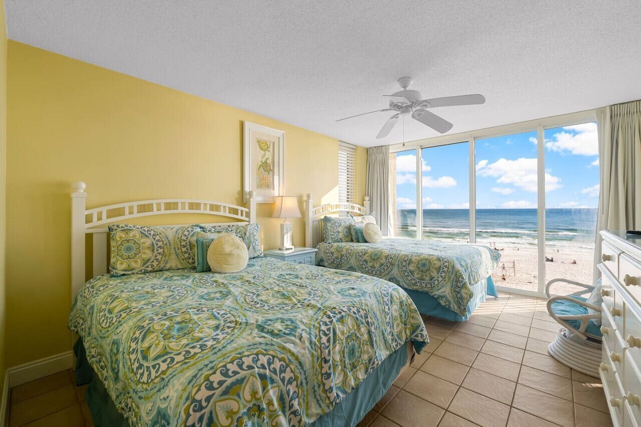 Un dormitorio con dos camas, un ventilador de techo y un ventanal con vista a la playa.
