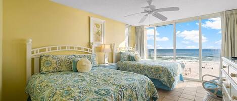 Un dormitorio con dos camas, un ventilador de techo y un ventanal con vista a la playa.