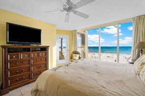 A bedroom with a large bed, a television on a wooden dresser, and a view of the beach.