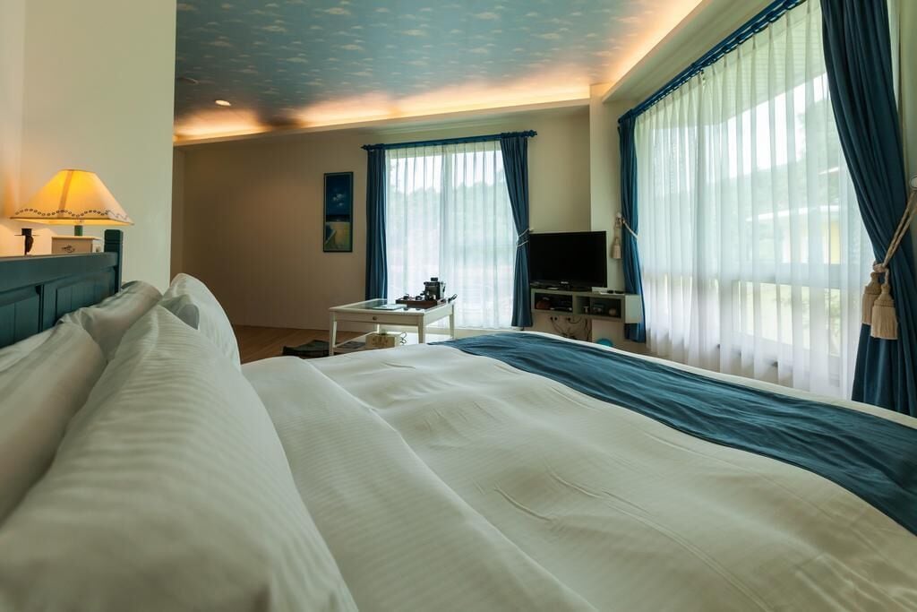 Quarto de hotel com uma cama grande, uma escrivaninha com abajur, televisão e cortinas nas janelas.