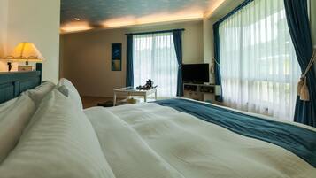 Quarto de hotel com uma cama grande, uma escrivaninha com abajur, televisão e cortinas nas janelas.