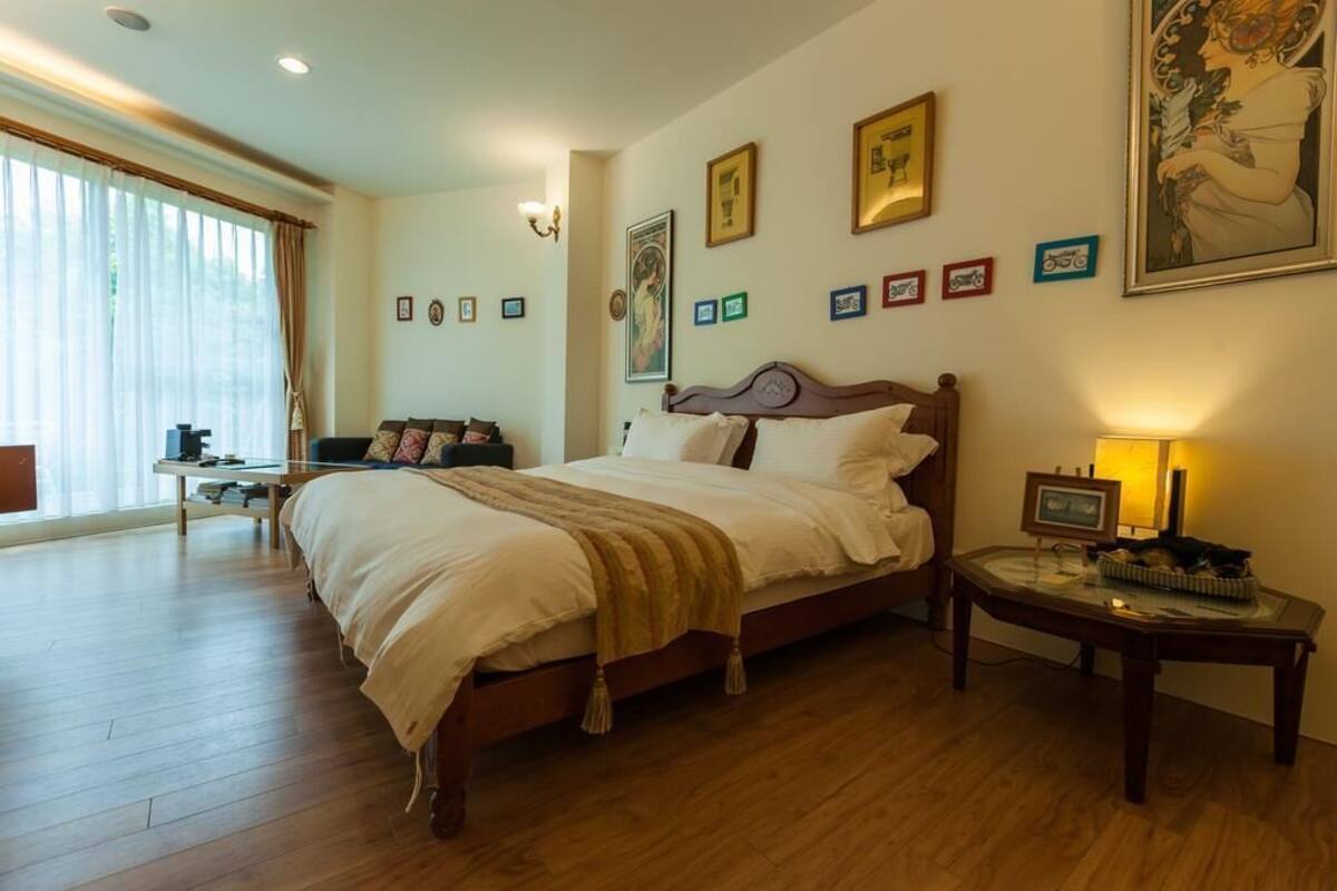 A bedroom with a wooden bed, a bedside table with a lamp, and a large window with curtains.