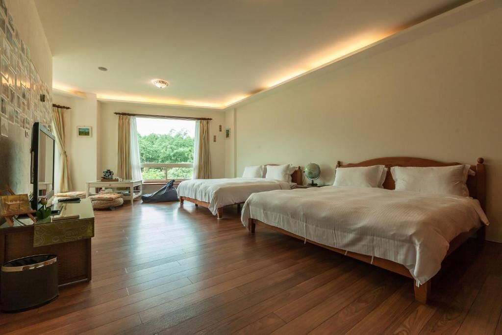 A hotel room with two beds, a desk, a TV, and a window with a view of greenery.