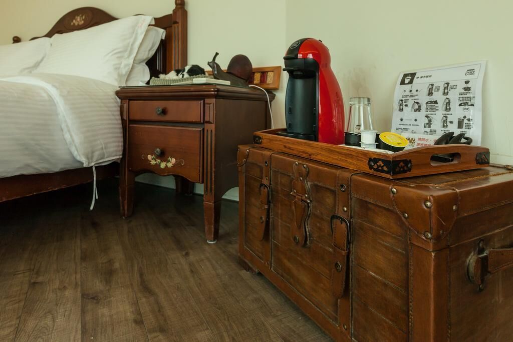 A wooden bed with white linens, a small wooden nightstand with a lamp, and a vintage suitcase on a wooden dresser.